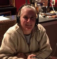 Laura Andrews, middle aged woman wearing fleecy sweatshirt and headset holding eyeglasses while seated at a table with a laptop in a room with burgundy wallls and wooden shelves. 
